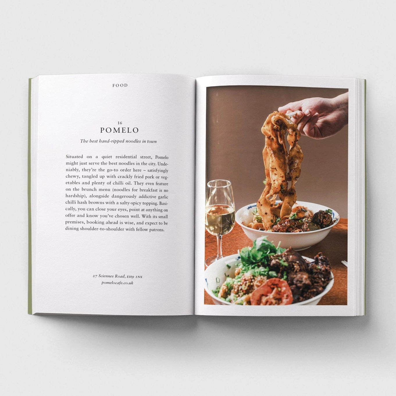 An open printed cookbook or magazine spread laid flat on a light gray background. The left page contains a recipe titled 'POMELO' with text, and the right page shows a food photograph of pasta being stretched with a glass of white wine and a plated dish below.