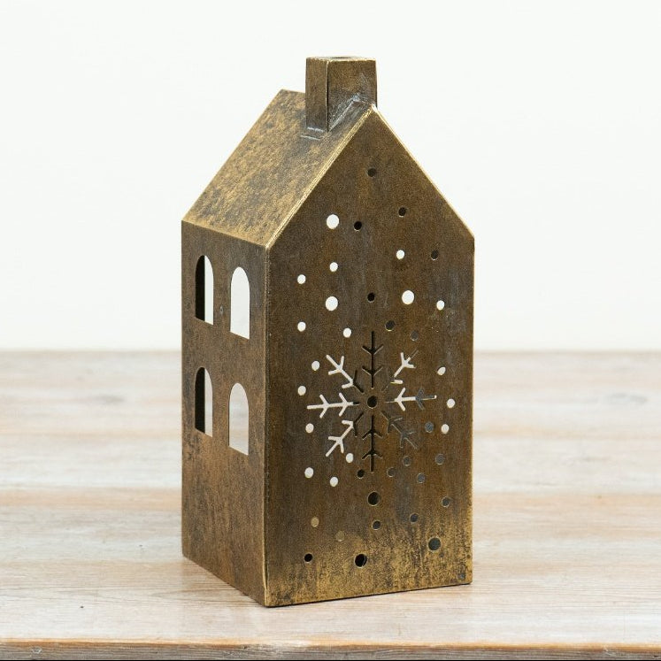 A small house-shaped decorative candle lantern in a distressed bronze finish, featuring laser-cut windows, a snowflake design on the front, and ventilation holes, standing on a light wooden surface against a white background.