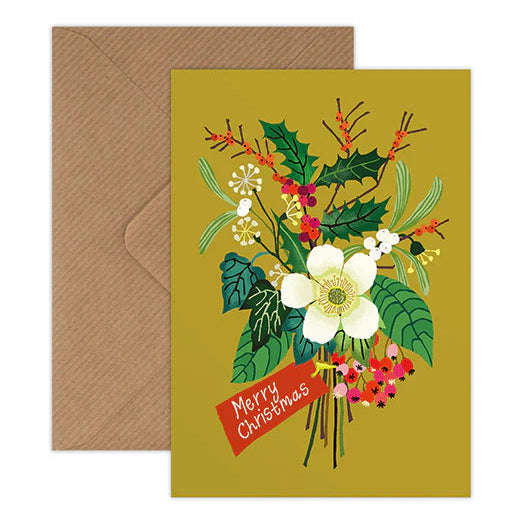 A square Christmas greeting card with a floral illustration on a mustard yellow background, shown with a brown envelope behind it on a white studio background. The card features white flowers, green leaves, red berries, and a red banner reading 'Merry Christmas'.