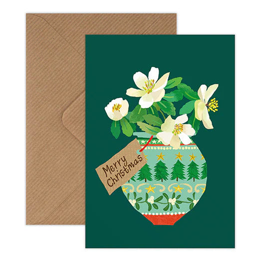 A Christmas greeting card with a dark green background, featuring an illustration of white flowers in a patterned green and red pot, shown with a brown envelope behind it.