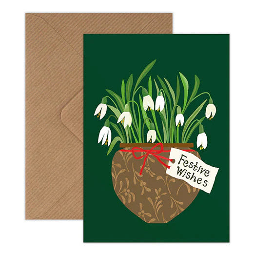 A square greeting card with a dark green background featuring an illustration of white snowdrops in a brown patterned pot tied with a red ribbon and a small tag reading 'Festive Wishes'. A plain brown envelope is placed behind the card on a white background.