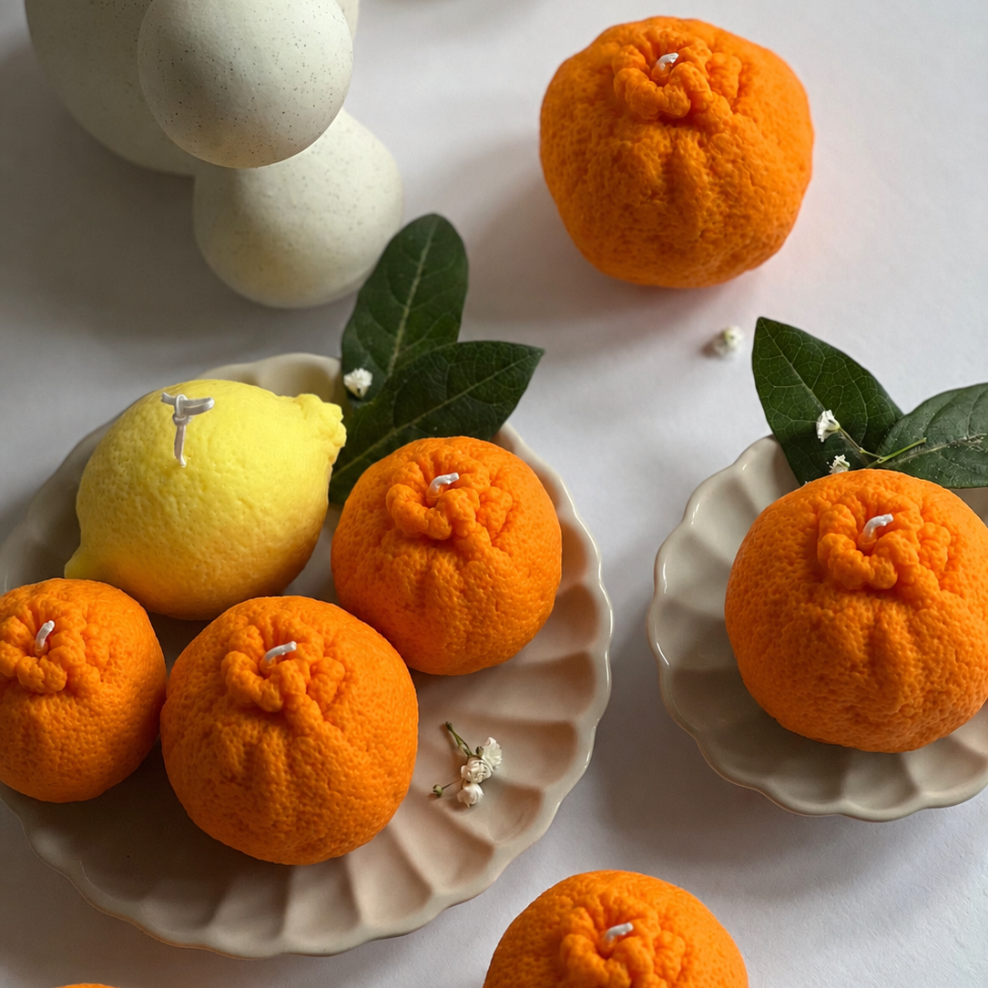 A styled flat-lay of orange pom-pom candles shaped like oranges, arranged on beige ceramic plates with a yellow pom-pom candle shaped like a lemon, green leaves, and neutral decorative props on a light background.