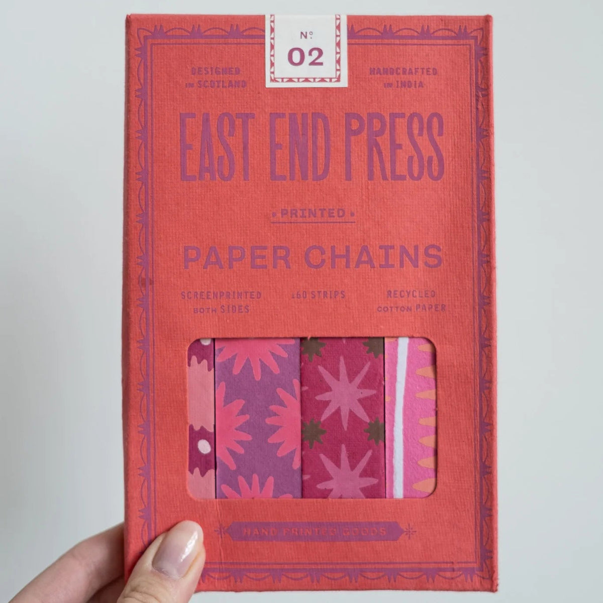 A hand holding a coral red package of East End Press Paper Chains, showing the colorful patterned paper chains visible through a clear window on the front.