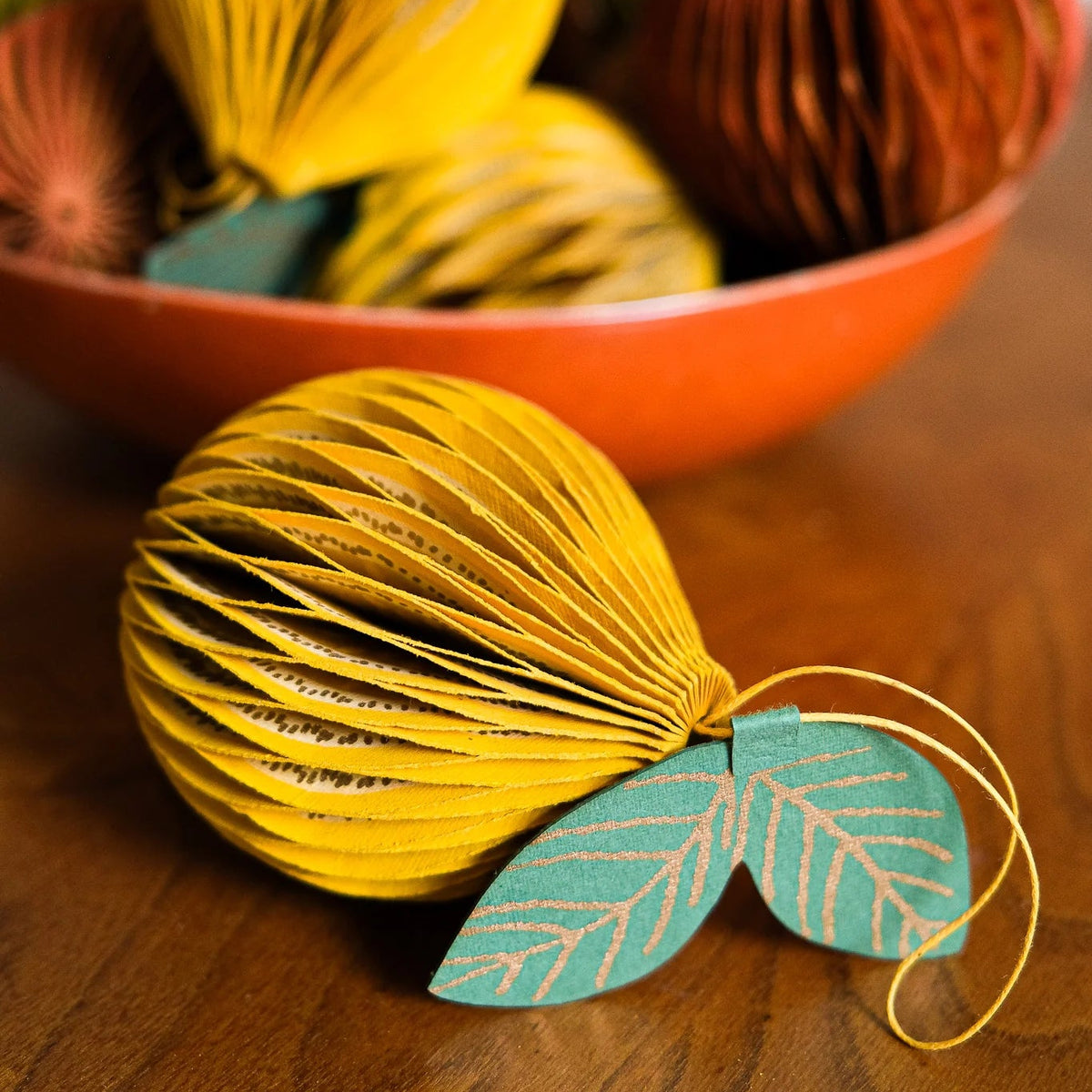 A decorative paper lantern ornament in bright yellow sits on a wooden table in the foreground, attached to two teal leaves. A matching orange bowl filled with other colorful paper lanterns is blurred in the background.