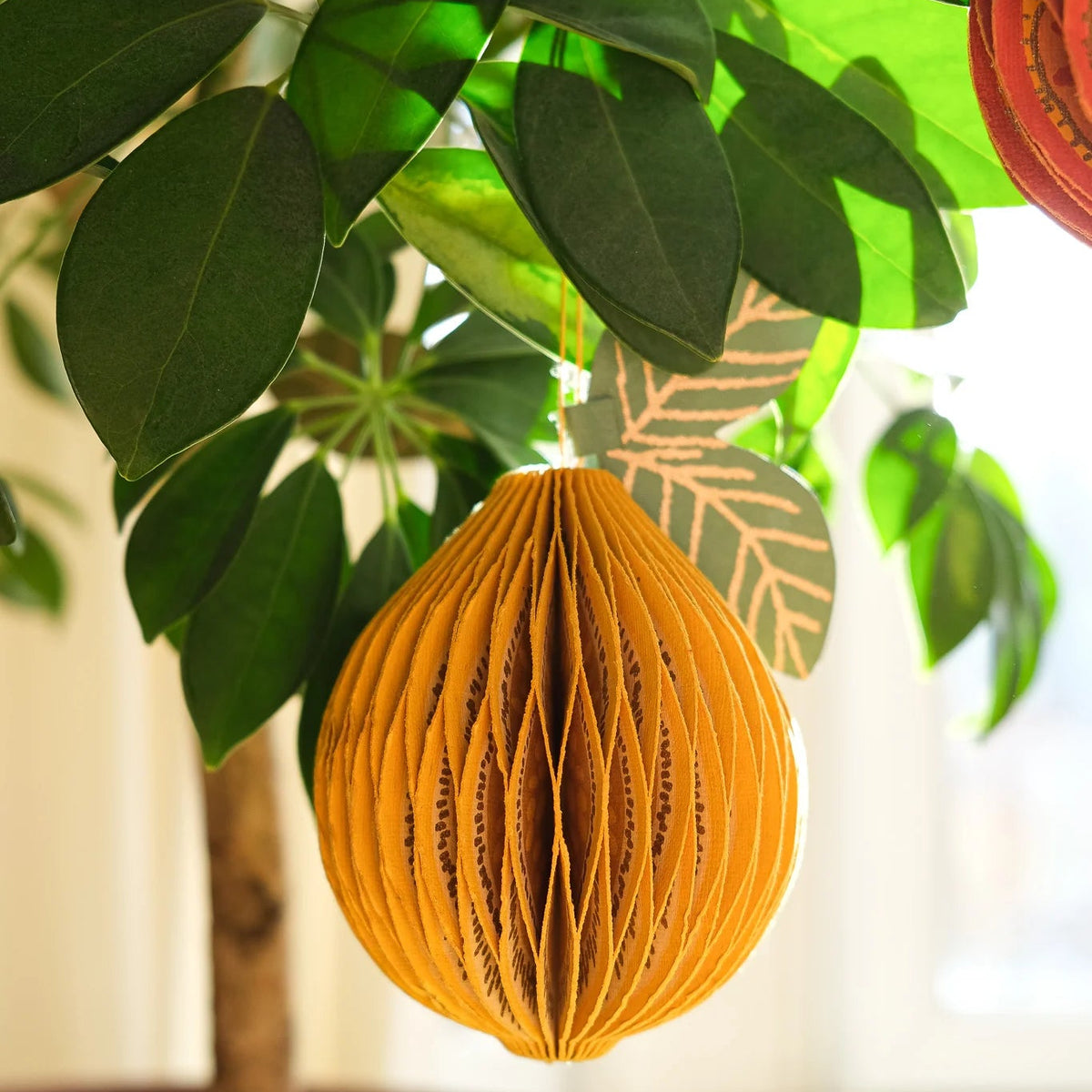 A golden-orange honeycomb paper hanging ornament shaped like a teardrop, suspended from green leaves on a potted plant in a bright indoor setting.