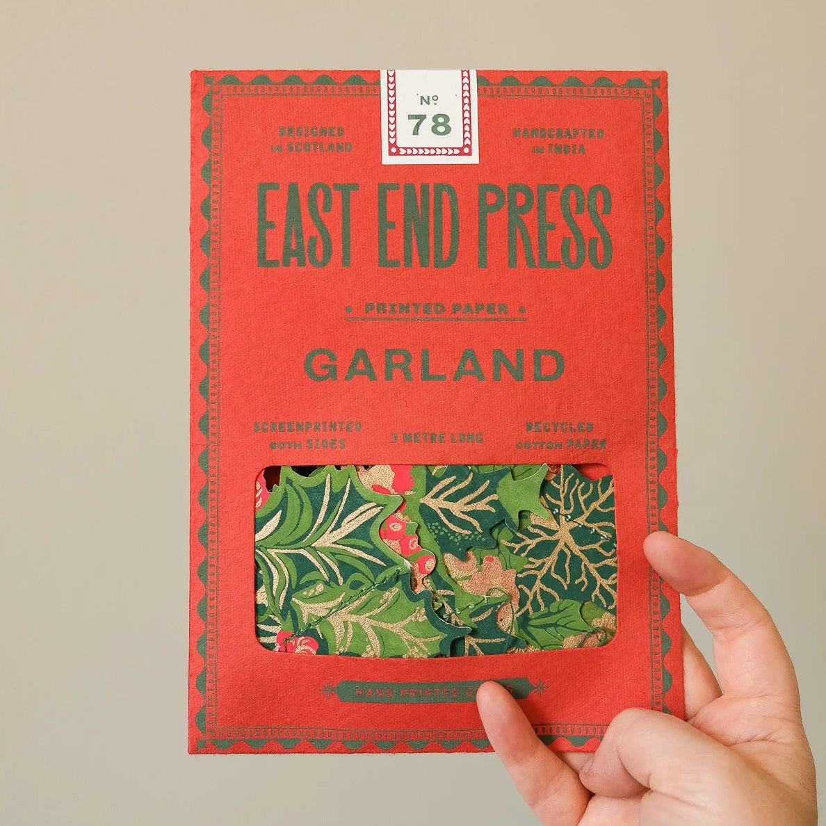 A hand holding a red East End Press Garland paper pack, showing a window that reveals a green and red holly-patterned sheet of paper, against a plain beige background.