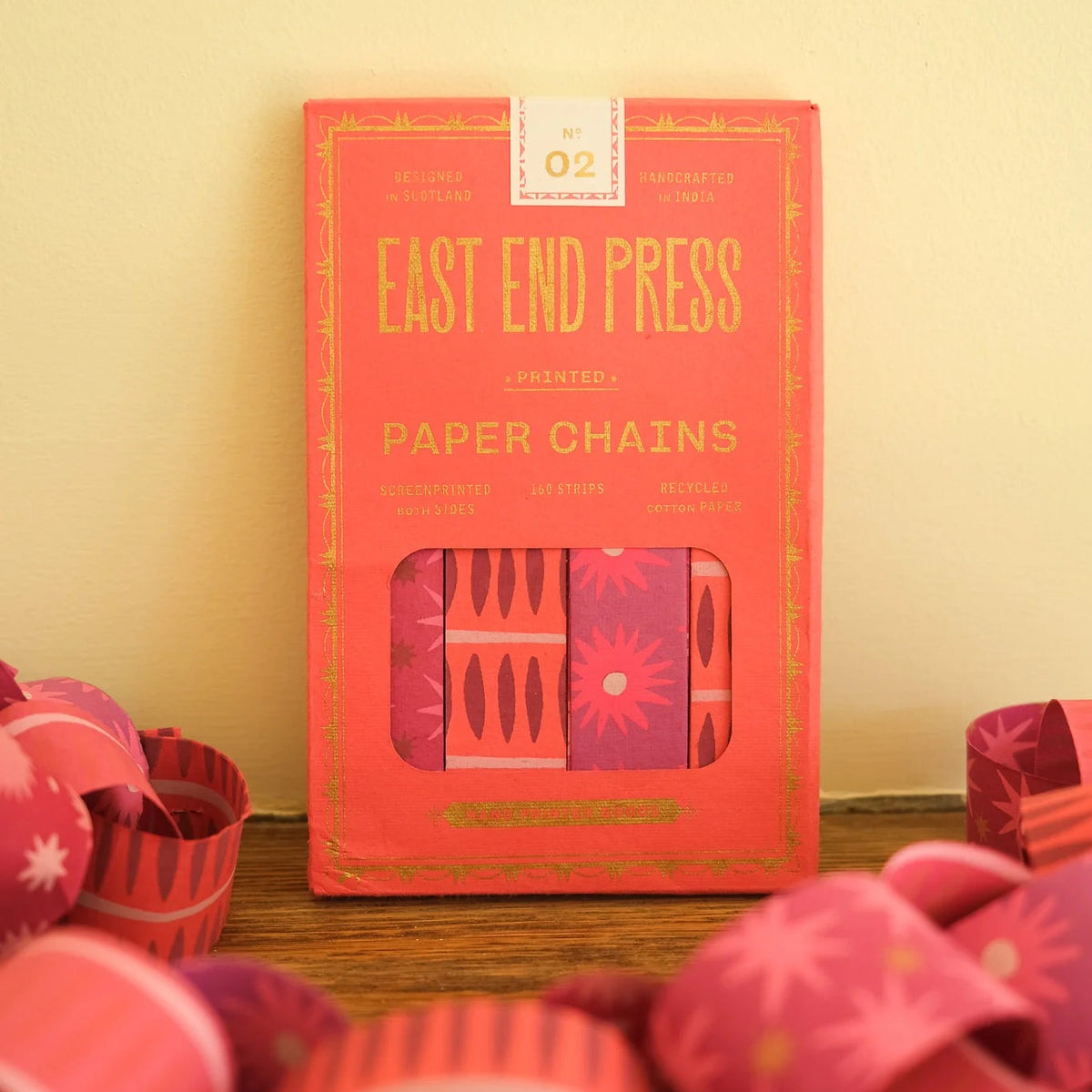 A box of East End Press paper chains in a coral red color, displayed on a wooden surface with pink and red paper chains scattered in the foreground against a cream-colored wall.