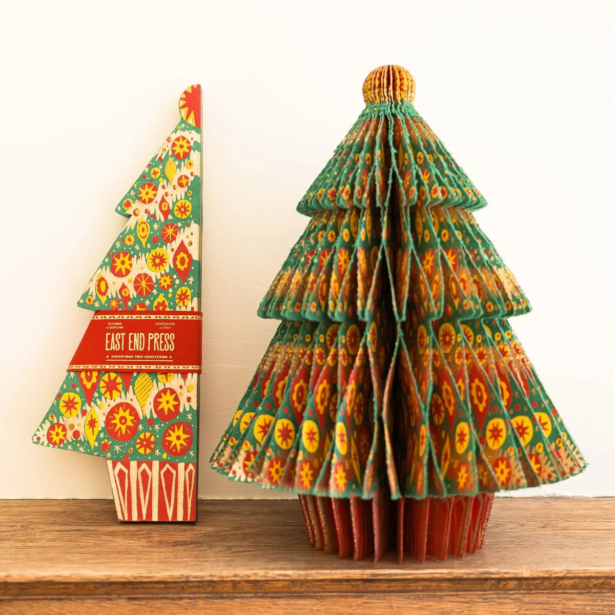 A decorative Christmas tree made from folded patterned paper stands on a wooden surface against a plain white wall. A matching paper tree box labeled 'EAST END PAPER' sits beside it.