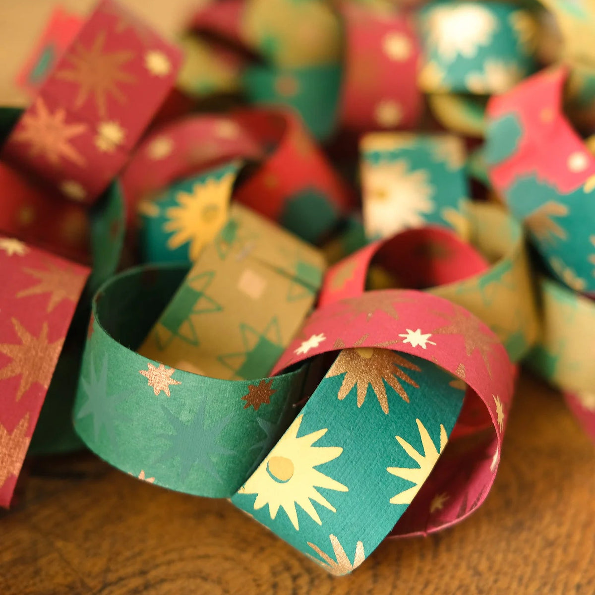 A close-up of a handmade paper chain garland with alternating red and teal patterned loops, resting on a wooden surface.