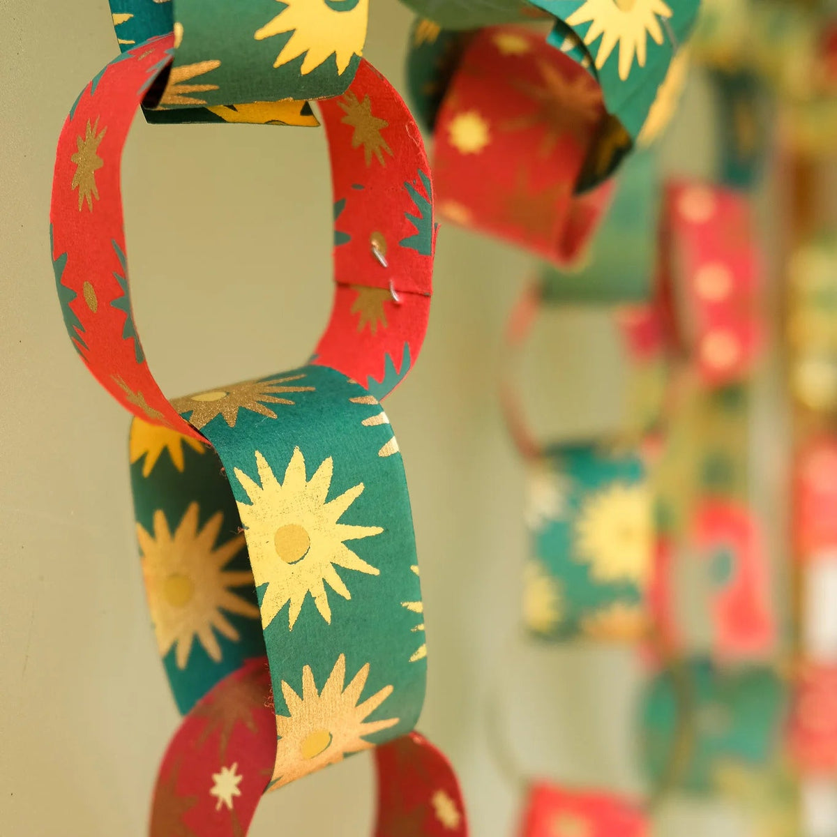 A close-up of a hanging paper chain garland with alternating red and green loops printed with yellow sunburst and star patterns, photographed against a softly blurred indoor background.