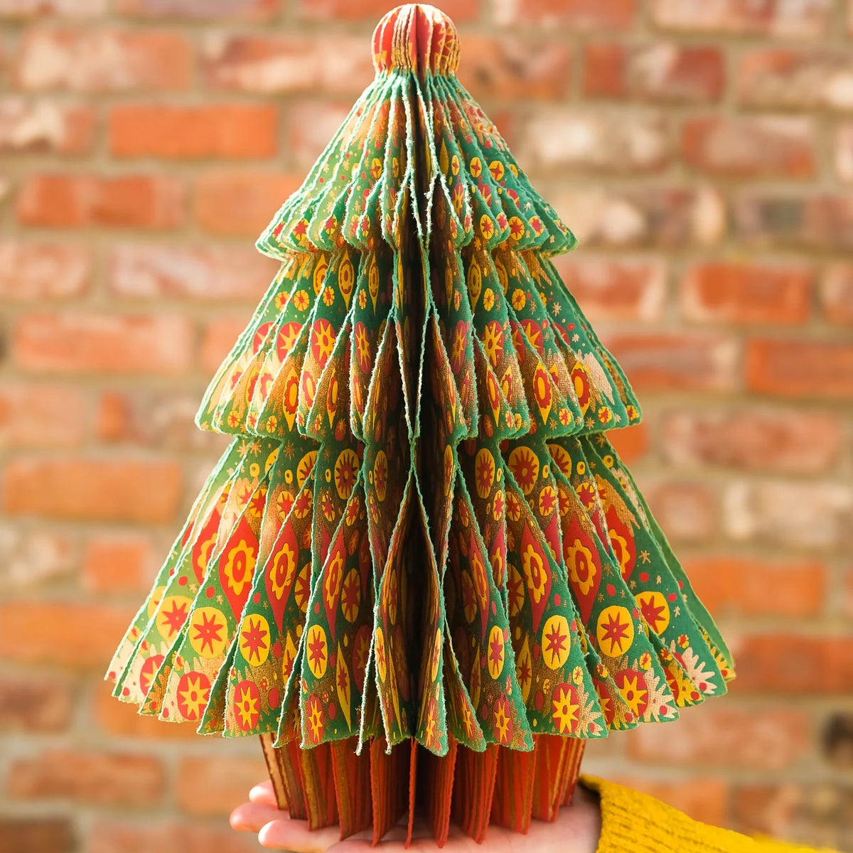 A hand wearing a yellow knit sweater holds a decorative, tiered Christmas tree sculpture made from patterned green paper. The background is a blurred red brick wall.