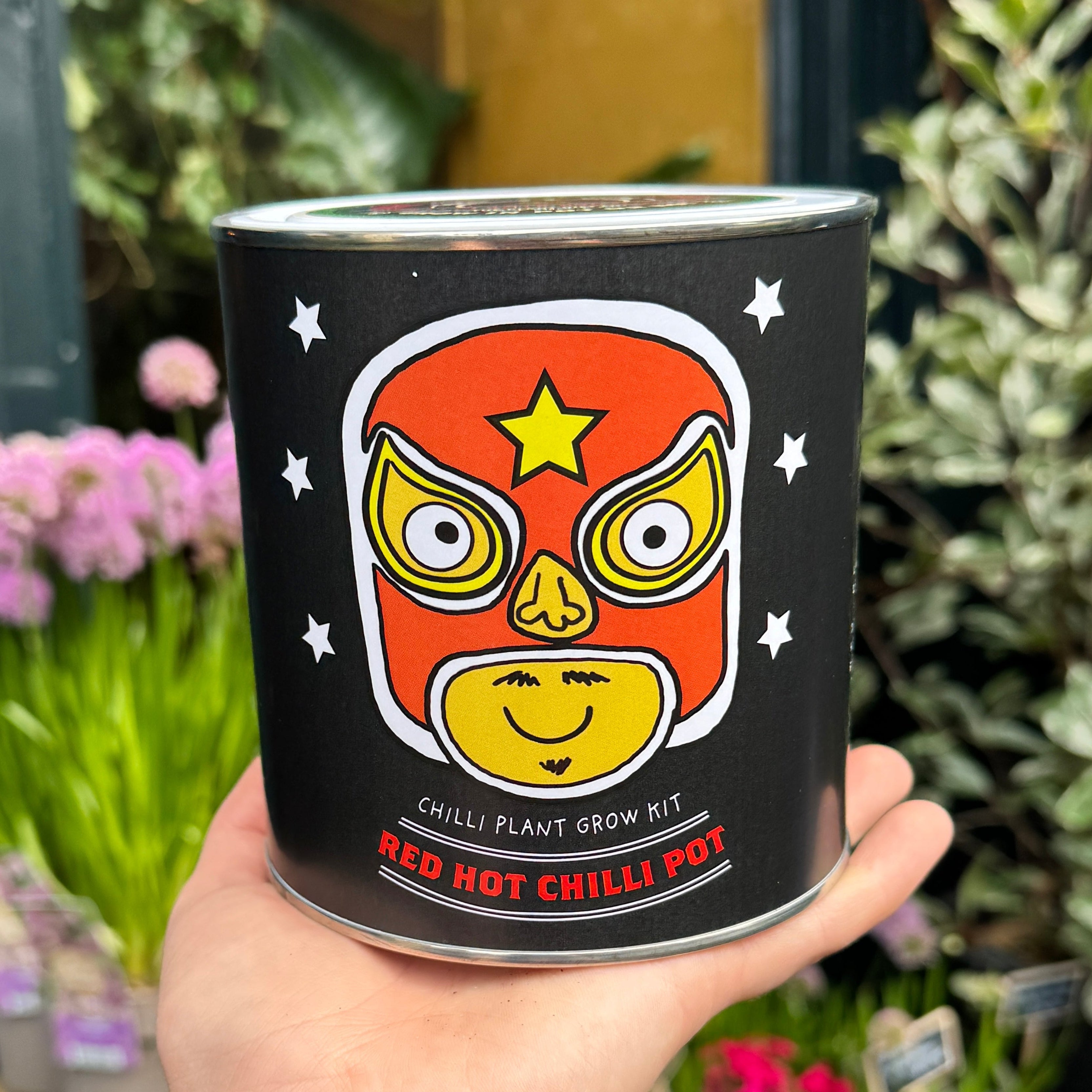 A hand holds a black cylindrical canister labeled 'Chilli Plant Grow Kit' and 'Red Hot Chilli Pot' with a colorful luchador mask illustration, set against a blurred garden background.