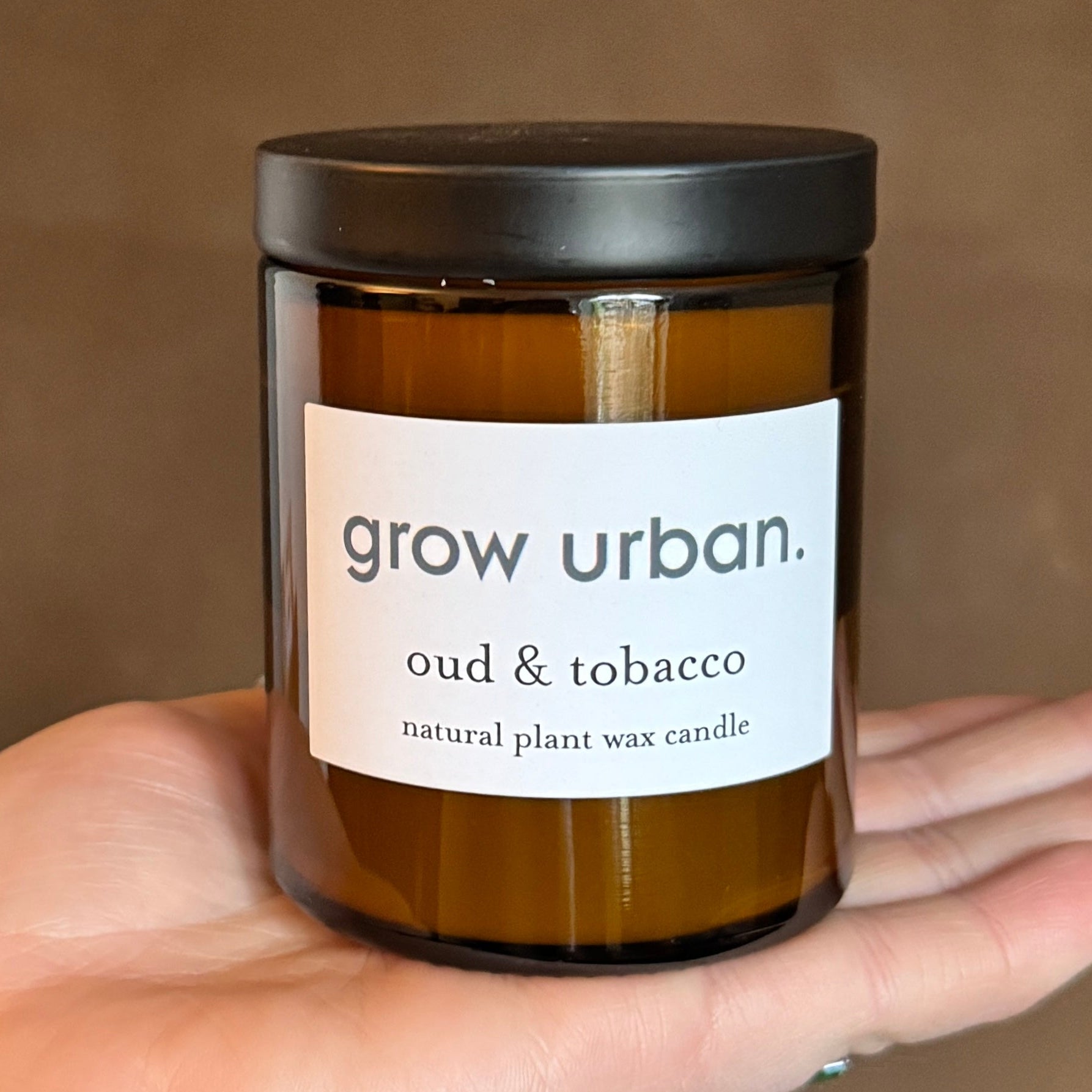 Candle jar labeled 'grow urban' with 'oud & tobacco' scent held in a hand against a brown background