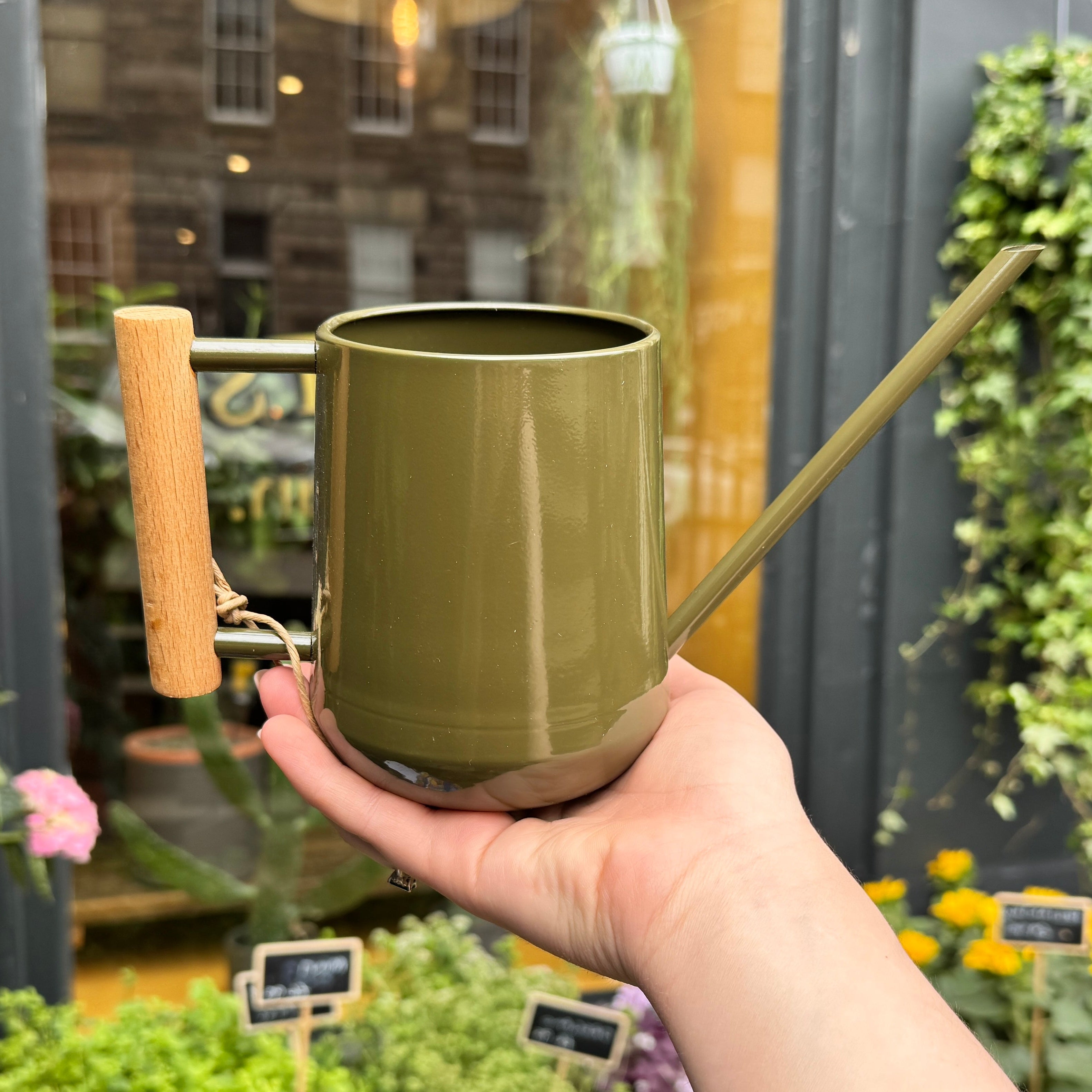 Hand holding an olive green watering can with a wooden handle in an outdoor setting with plants.