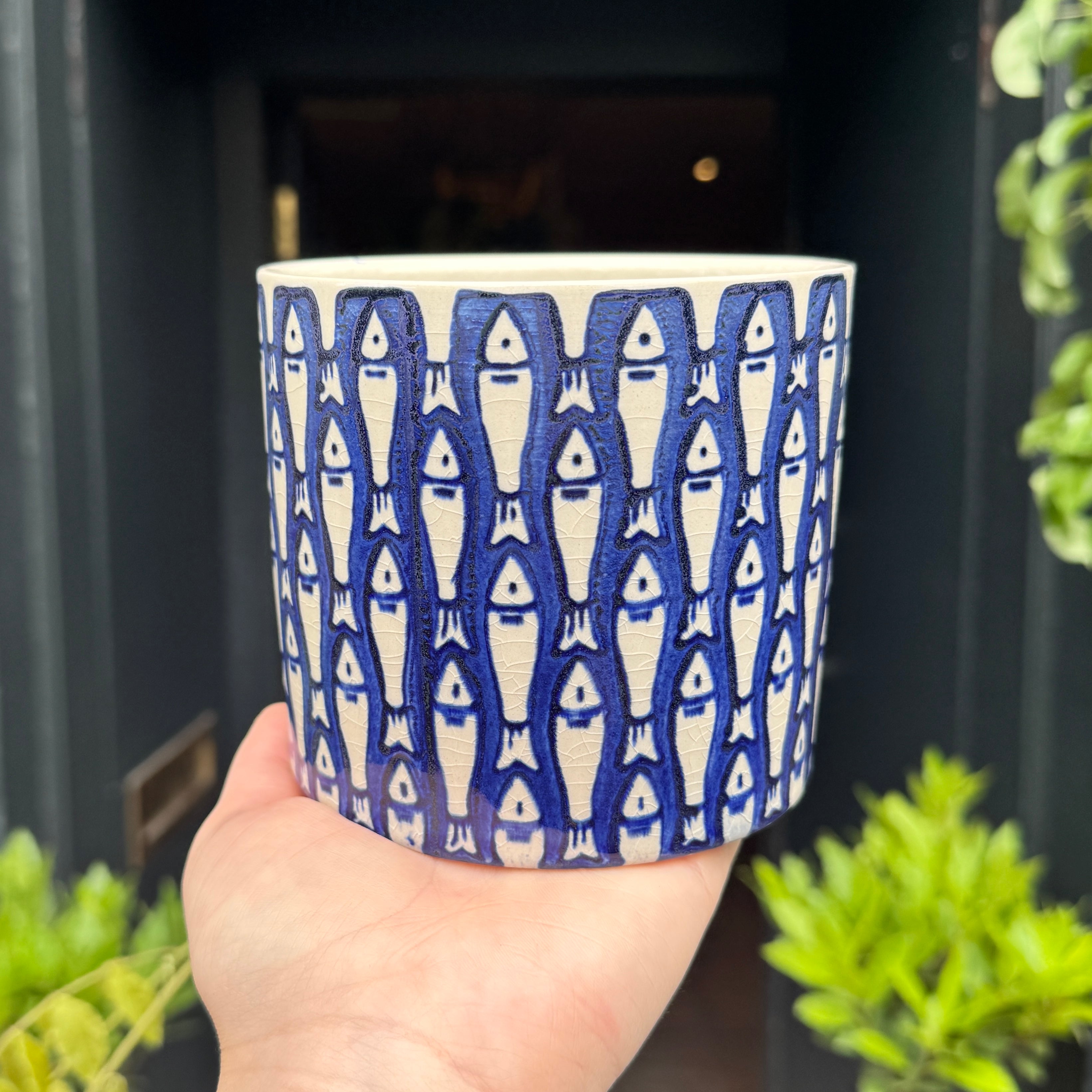 Hand holding a blue and white fish patterned plant pot with greenery in the background