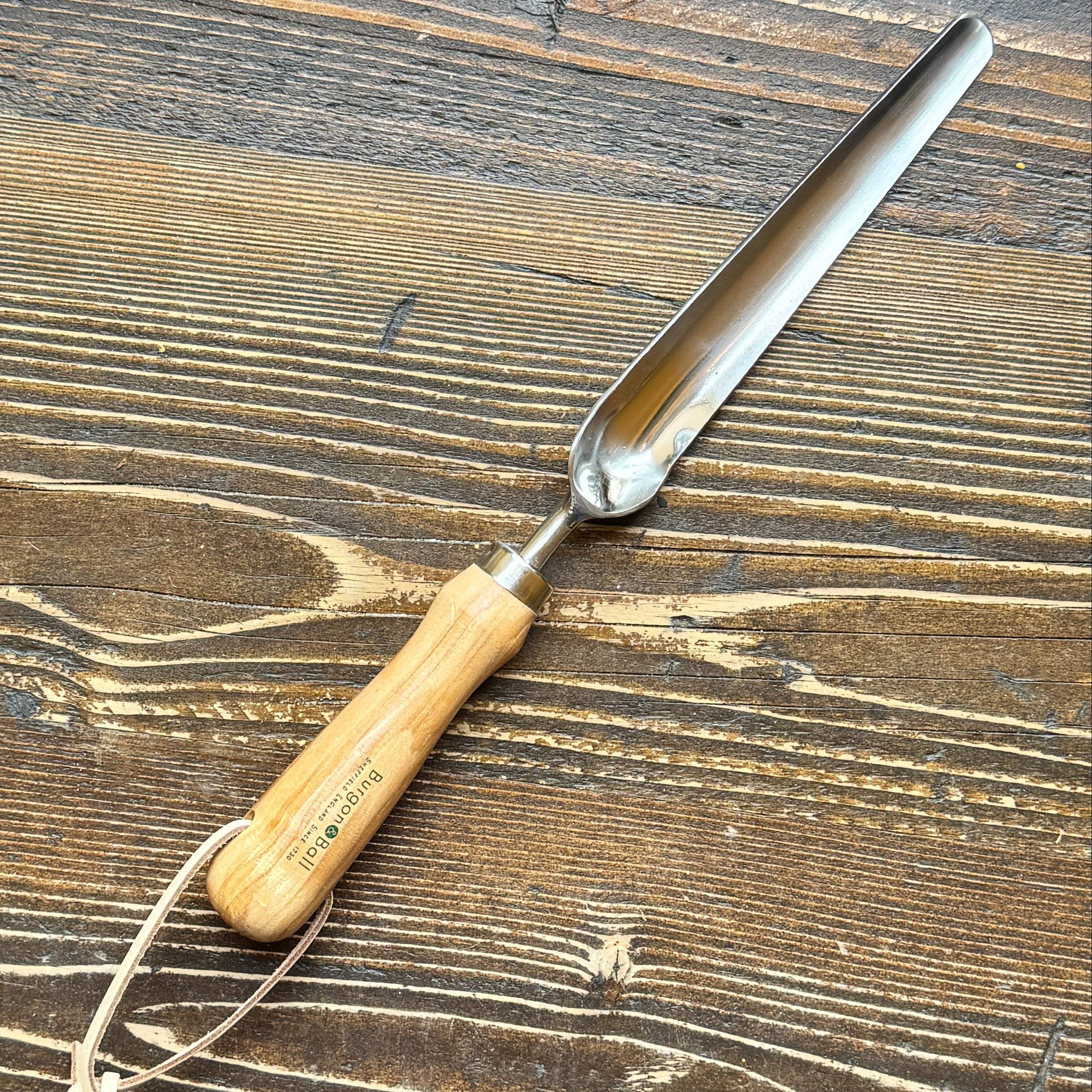 A metal garden widger with a light wood handle and a white hanging string, placed diagonally on a dark brown wooden surface.