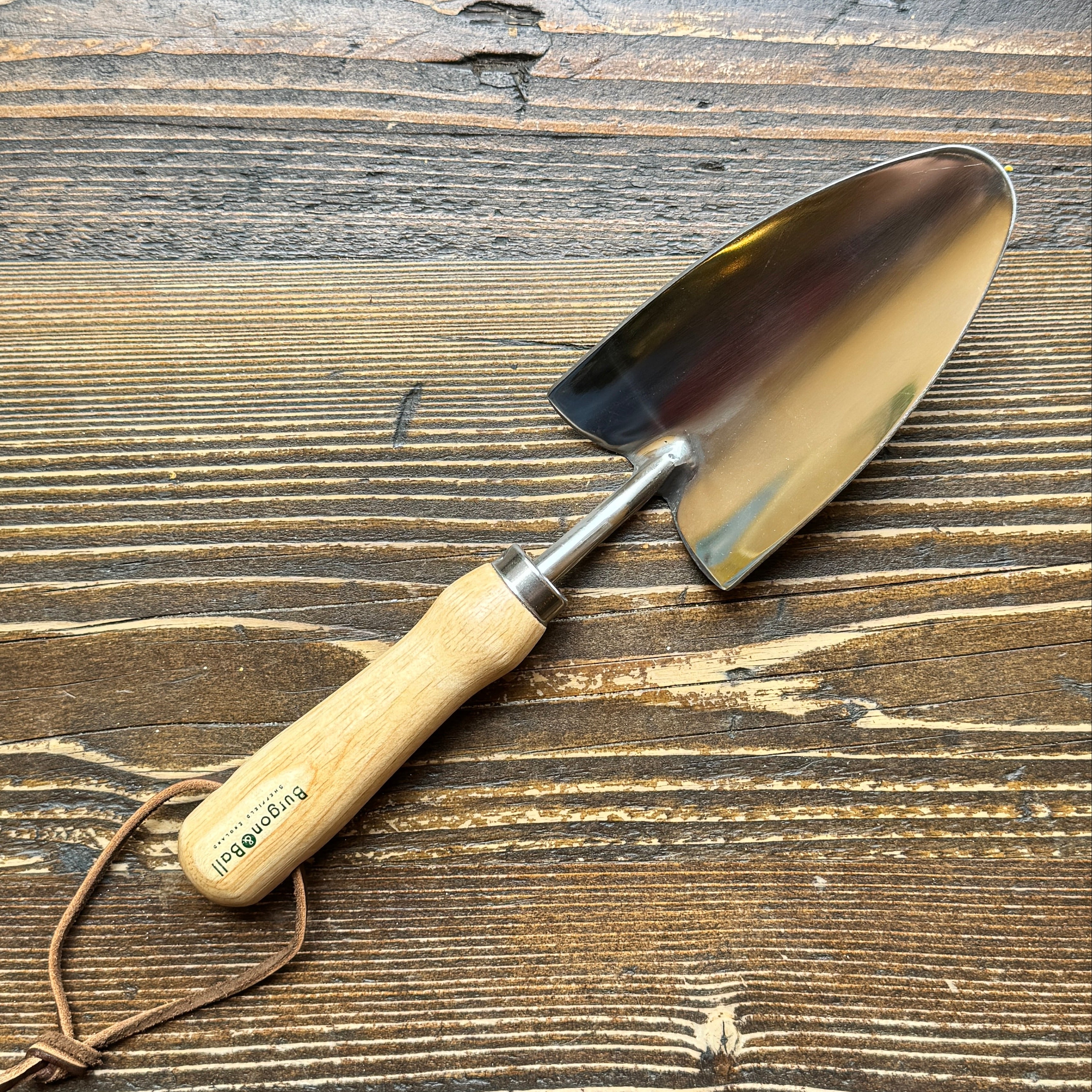 A small hand trowel with a light wood handle and a polished steel blade, resting on a rustic dark wood surface. The handle is printed with the brand name 'Burgon & Ball'.