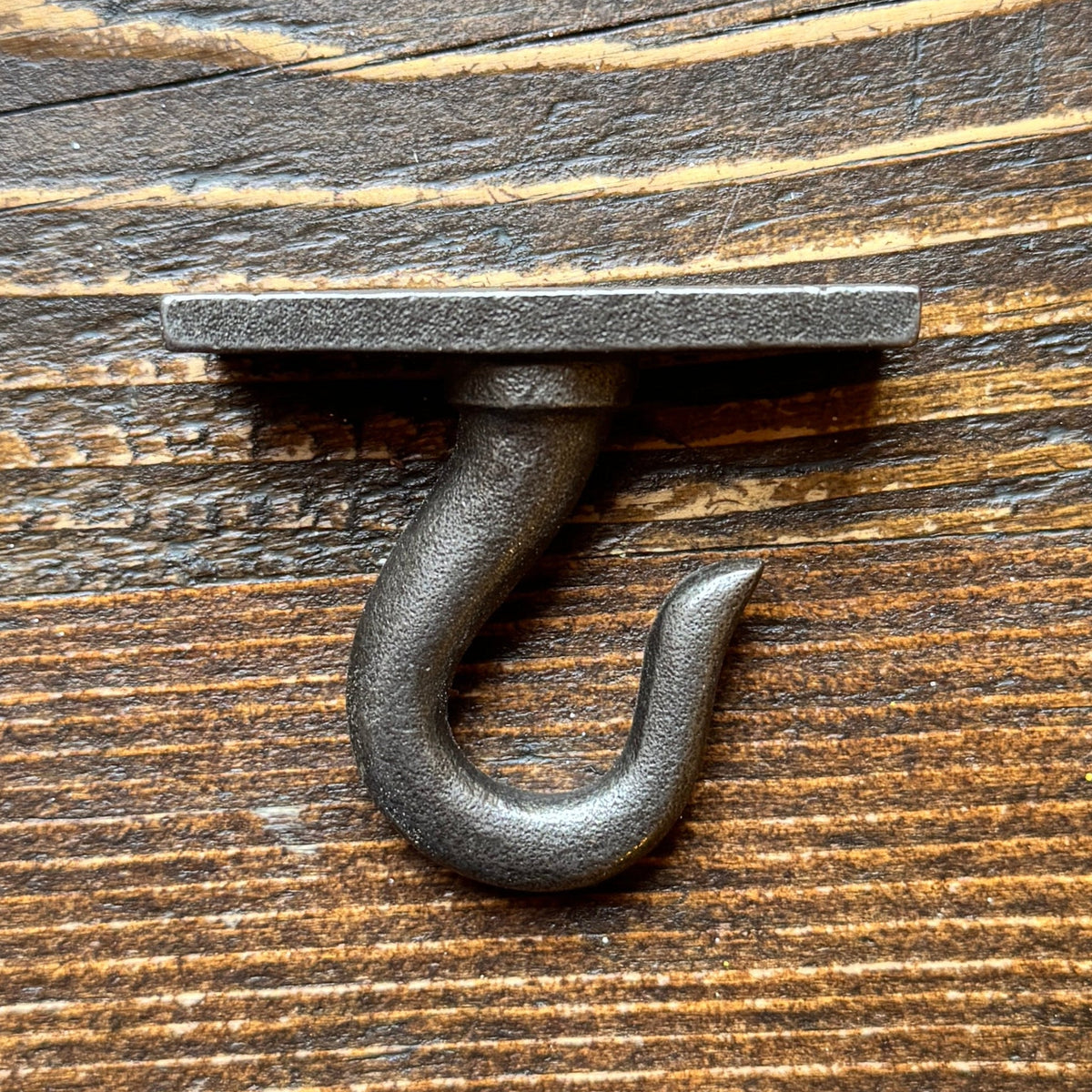 A rustic cast iron hook with a flat mounting plate, shown against a dark brown wood grain background.