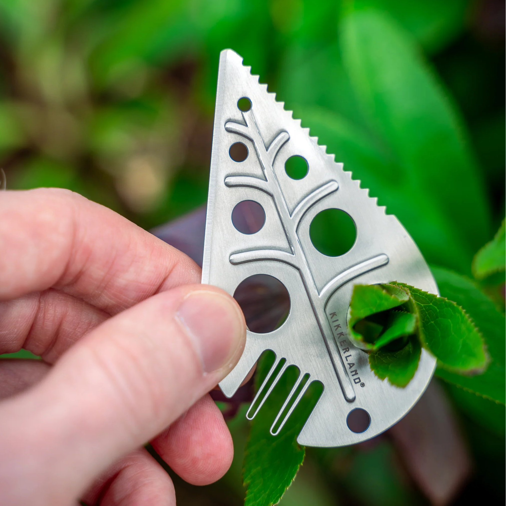 Hand holding a silver multitool with leaf design against a green leafy background