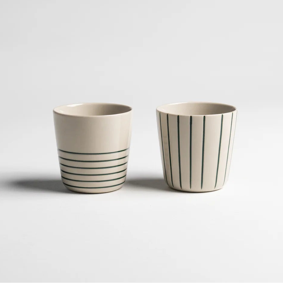 Two small ceramic cups on a light gray background, one with horizontal dark green stripes and the other with vertical dark green stripes, both on a warm off-white base.
