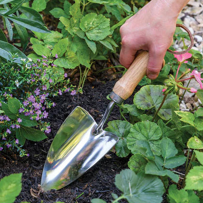 Hand using a garden trowel in a garden setting with plants and flowers.