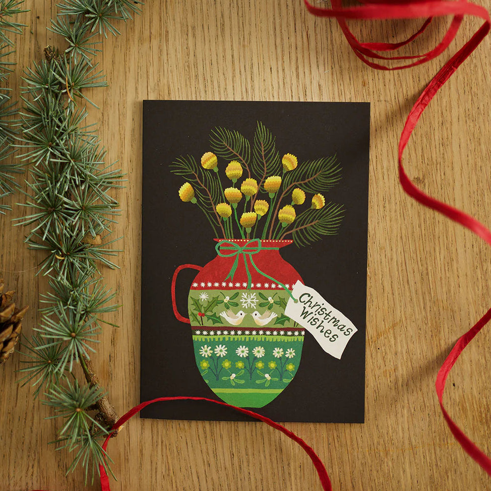 A square greeting card with a winter floral illustration, styled on a wooden surface with pine branches, a pinecone, red ribbon, and a spool of red ribbon.