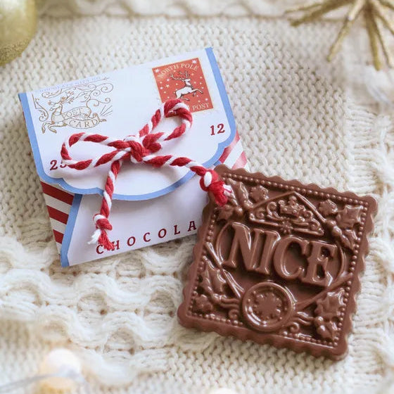 A square milk-chocolate bar embossed with the word 'NICE' in decorative lettering, placed beside a small wrapped chocolate gift box tied with red-and-white string on a cream knit blanket.