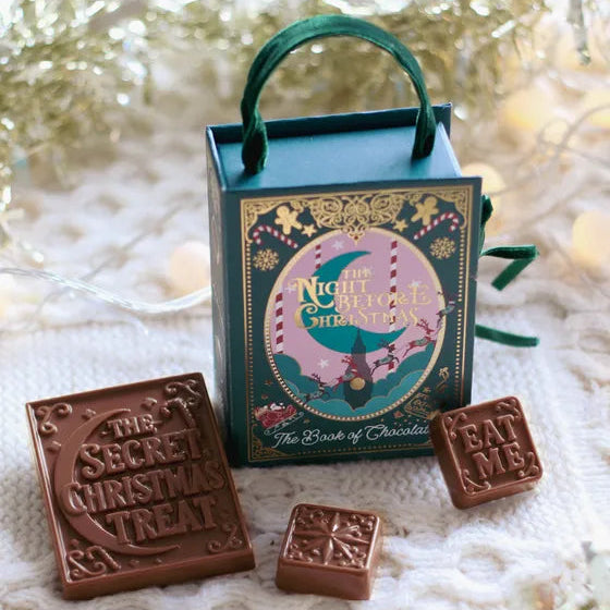 A small teal gift box with a green handle and decorative front label reading 'The Night Before Christmas' and 'The Book of Chocolates,' surrounded by three square milk-chocolate treats on a white knit surface with tinsel and warm bokeh in the background.