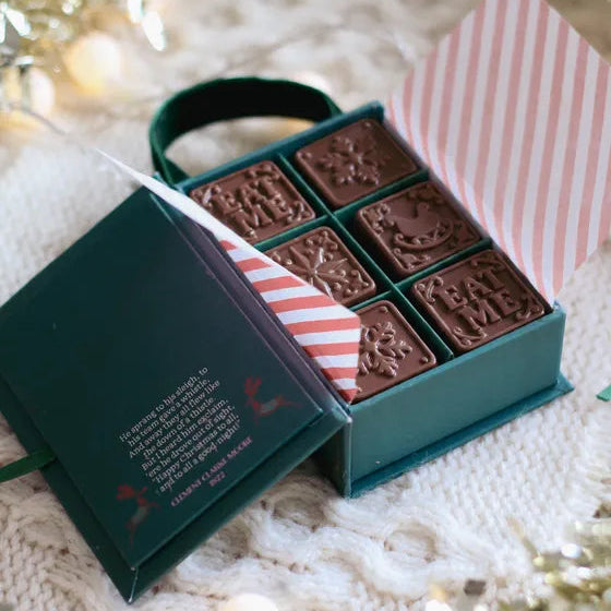 A dark green gift box of six chocolate truffles with holiday designs, shown open on a white knit blanket with festive lights in the background.