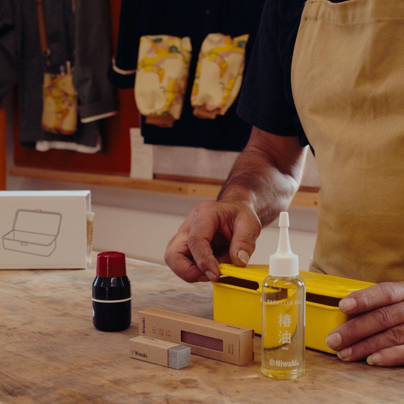 A person wearing a tan apron works at a wooden table, handling a Niwaki Tool Care Kit with Japanese garden tool cleaning and sharpening accessories.