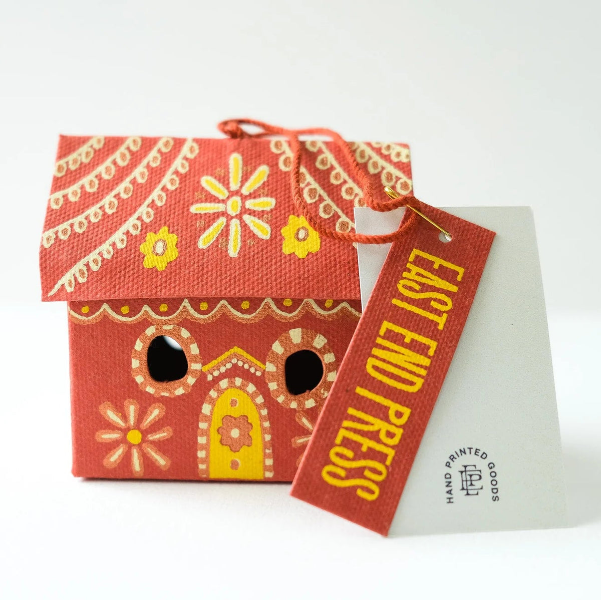 A small decorative house-shaped paper product in red-orange with yellow and white printed patterns, shown with a hanging tag that reads EAST END PRESS and a white backing card. No person, animal, or real environment is present, so this is Product/Packaging.