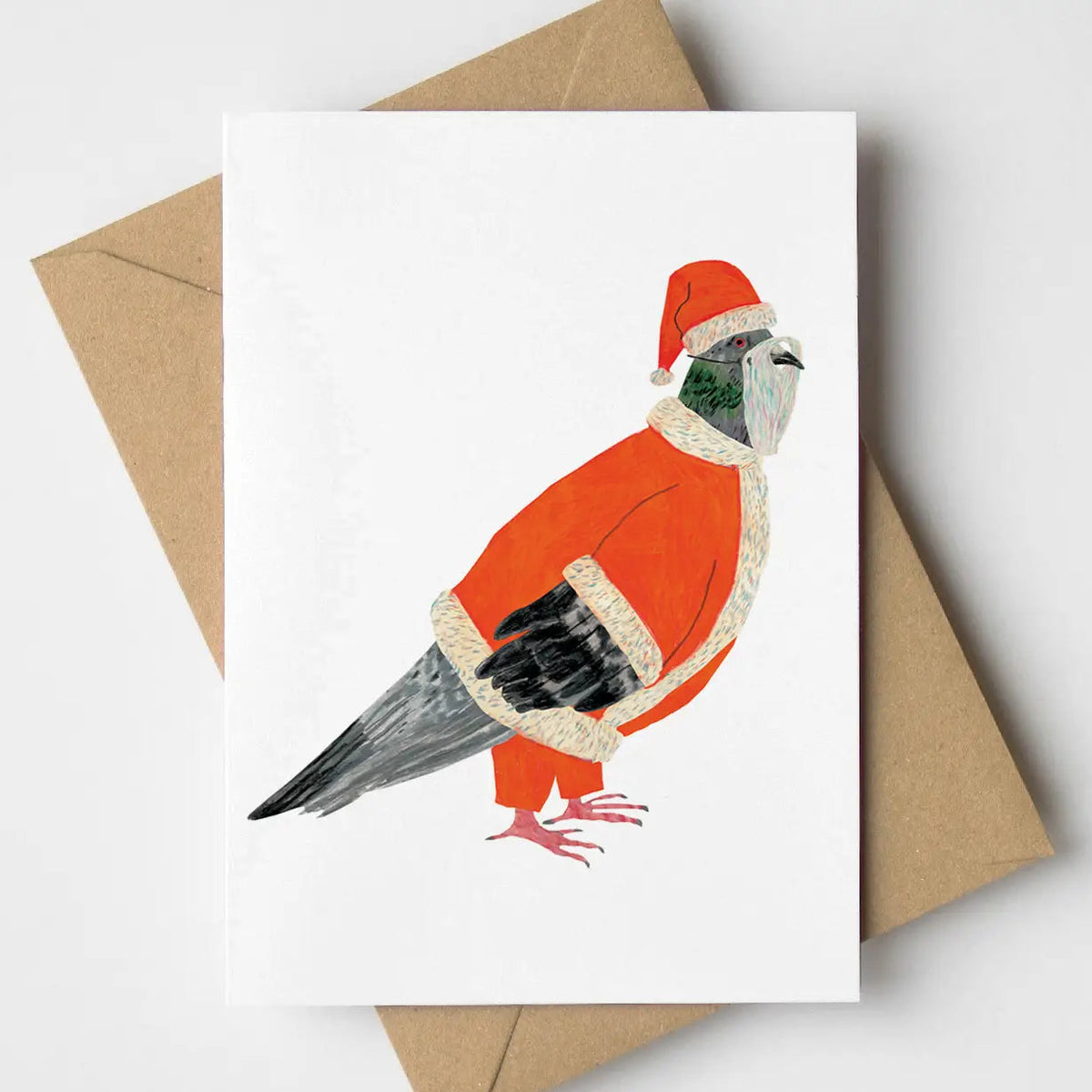 A white greeting card featuring a whimsical illustration of a pigeon dressed in an Santa-style coat with white trim and a red Santa hat, placed on top of brown kraft envelopes against a light gray background.