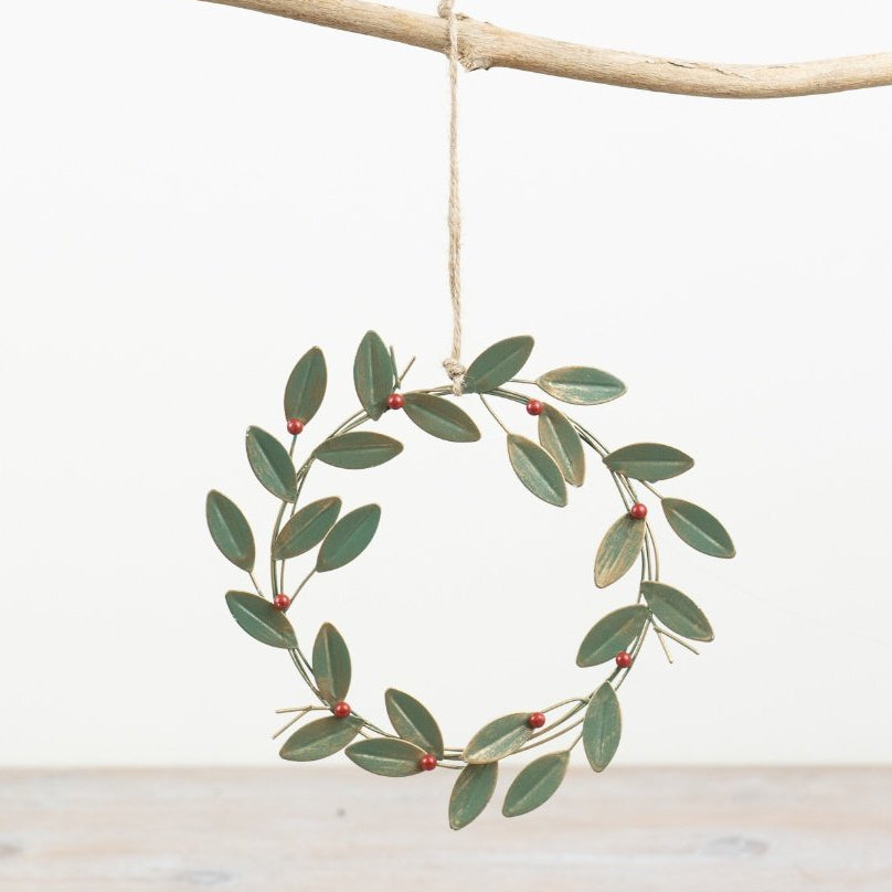 A small hanging wreath made of green leaves and tiny red berries, suspended from a natural branch against a clean white background with a light wood surface below.