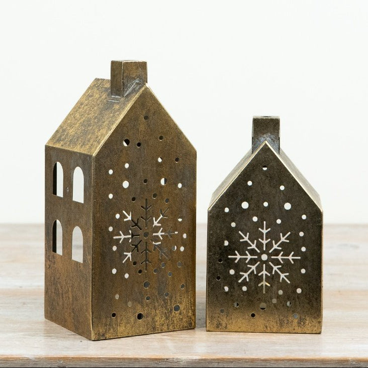 Two small house-shaped decorative lanterns in a distressed bronze finish, featuring snowflake cutouts, displayed on a light wooden surface against a white background.