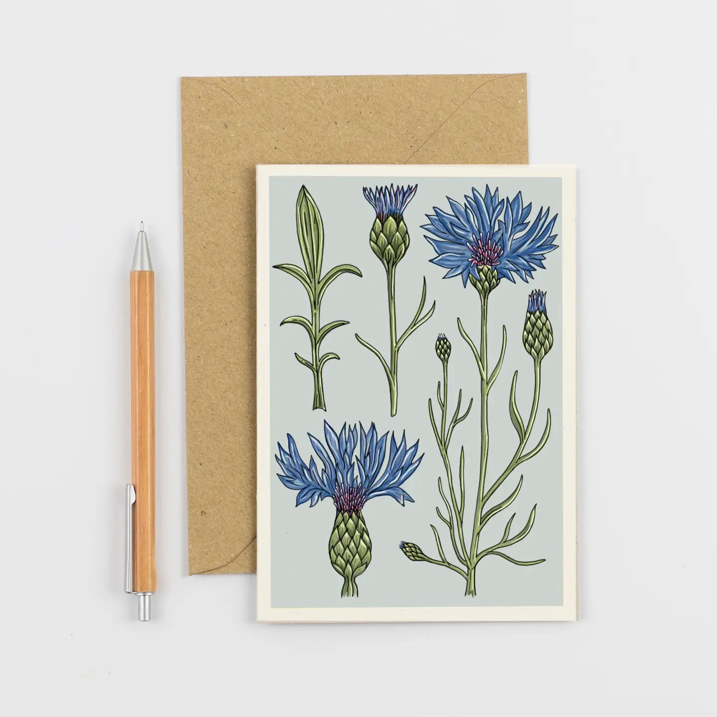 A botanical art print card featuring blue cornflowers on a pale blue-gray background, shown with a brown kraft envelope and a wooden pen on a light gray surface.