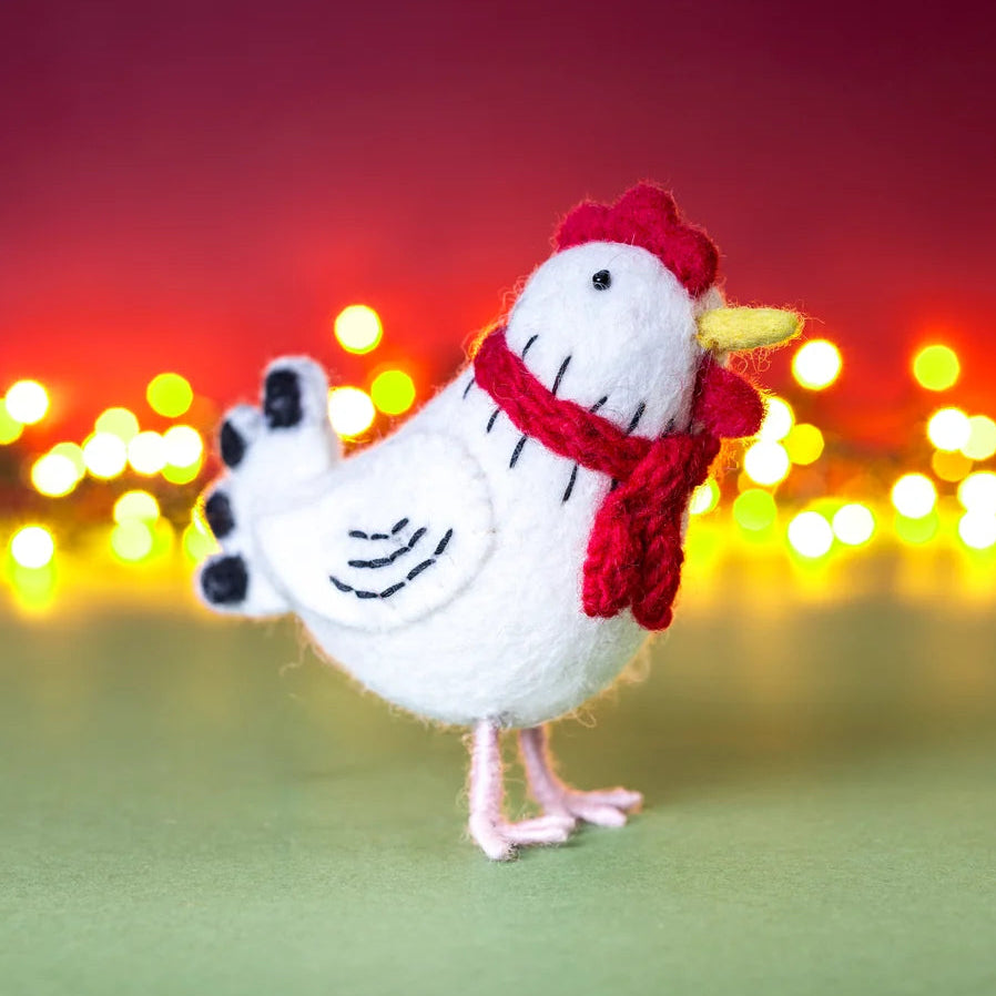 A small white felt chicken figurine wearing a red scarf and red comb, standing on a green surface with blurred warm lights in the background.