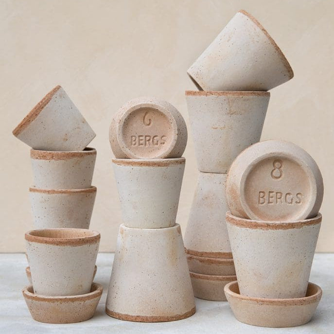 A set of unglazed ceramic plant pots in a warm sandy beige color, stacked in various heights and orientations on a light gray surface against a beige background. The bottoms of several pots are visible, showing embossed numbers (6 BERGS, 8 BERGS) and natural clay edges.