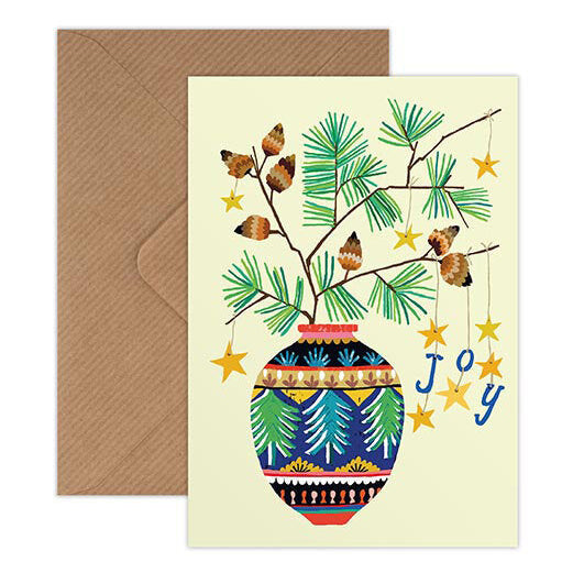 A greeting card with a brown envelope, featuring a festive illustration of a blue patterned vase with pine branches, pinecones, hanging gold stars, and the word 'joy' in blue script on a cream background.