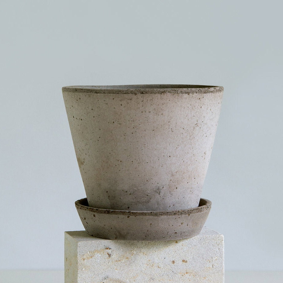 A speckled light grey Bergs Potter Juliepot with a matching saucer, displayed on a textured stone pedestal against a light grey background.