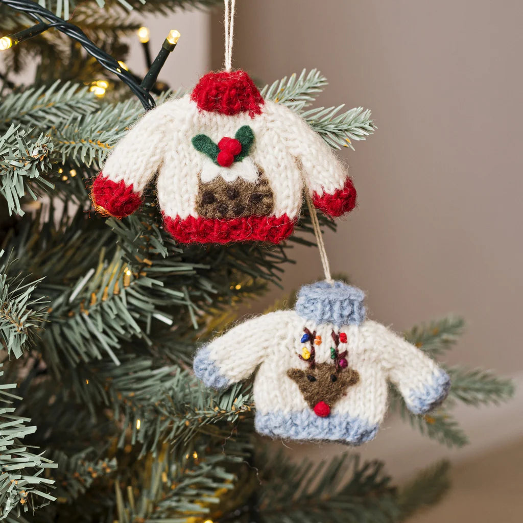 Two knitted Christmas sweater ornaments hanging on a decorated evergreen tree. The top ornament is cream with red trim, green holly, and a brown reindeer face.