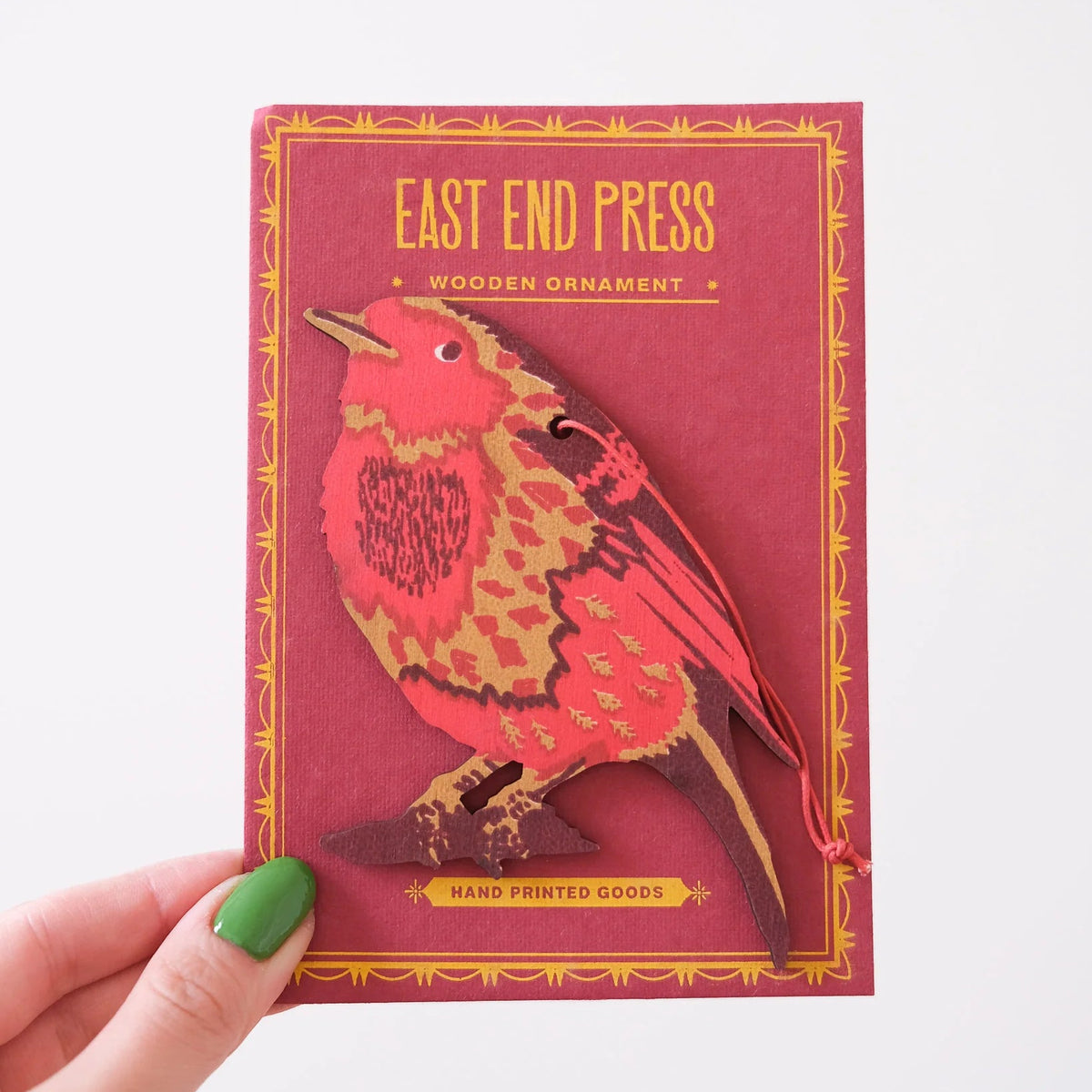 A hand with green nail polish holds a rectangular red paper packaging card for an 'East End Press' wooden ornament featuring a stylized Robin bird cutout.