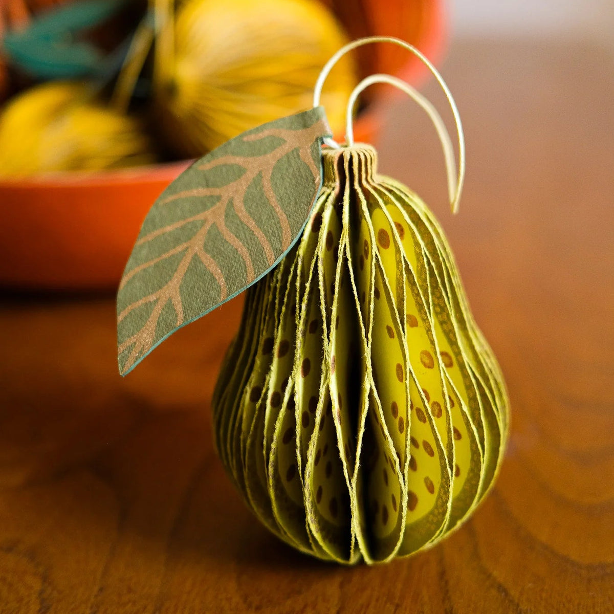 A decorative pear-shaped ornament made from layered yellow paper with a green leaf accent and a thin gold wire hanger, displayed on a wooden surface with a blurred background of similar ornaments.