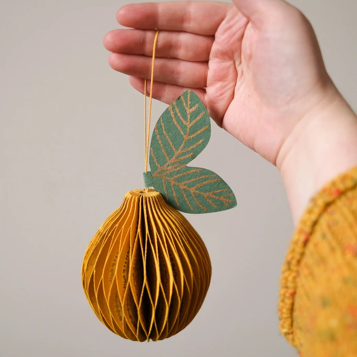 A hand holds a small hanging ornament shaped like a textured golden-yellow paper fruit with two green leaf accents, shown against a plain light gray background.
