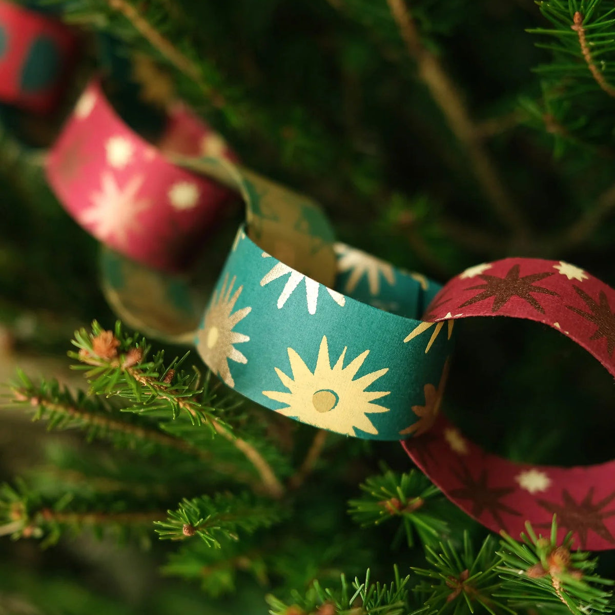 A close-up of a handmade paper garland made of linked loops in teal with white sunburst patterns and burgundy with white snowflake patterns, hanging on a green Christmas tree.