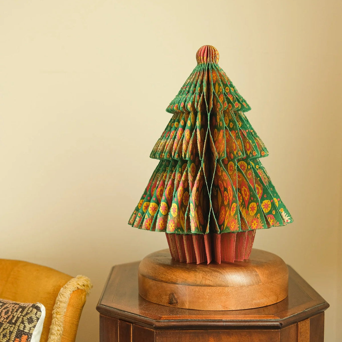 A decorative Christmas tree sculpture made from layered patterned paper or fabric, displayed on a round wooden base atop a dark wood side table against a beige wall.