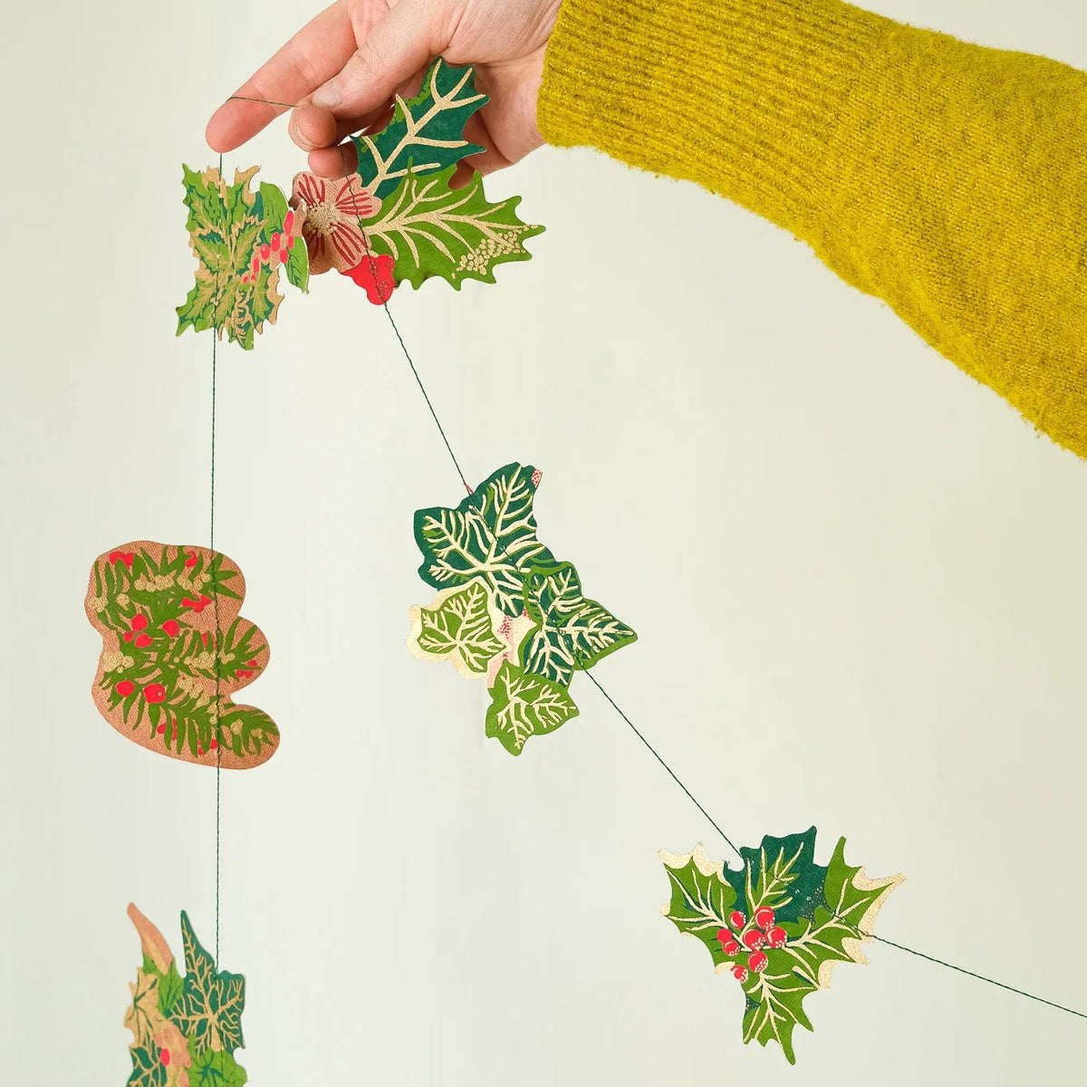 A hand wearing a yellow knit sweater holds a hanging Christmas garland made of green holly leaves and pinecones with small red berries against a plain light background.