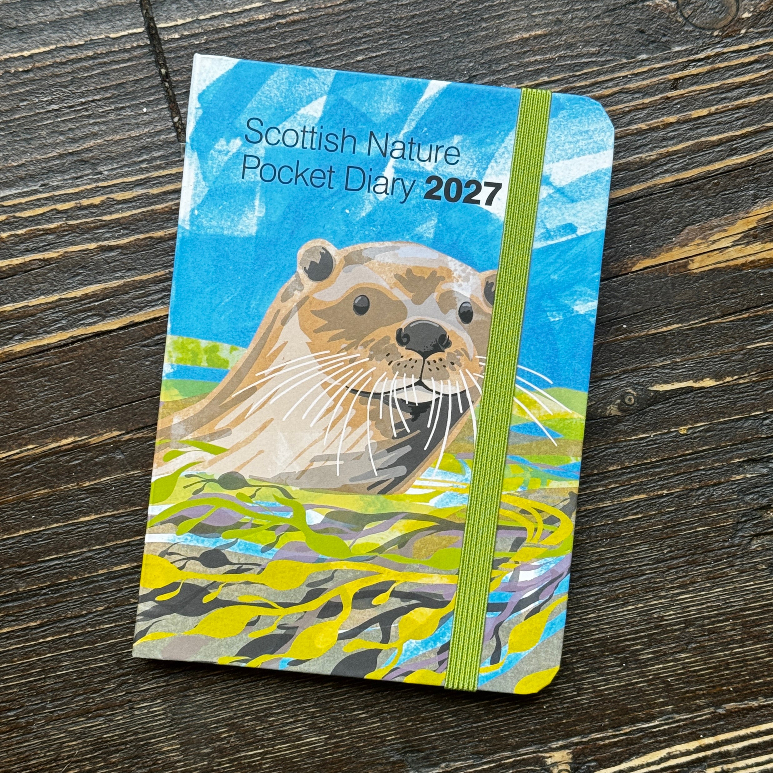 A pocket-sized 2027 Scottish Nature Diary featuring a colorful illustration of an otter on the cover, resting on a dark wood surface.