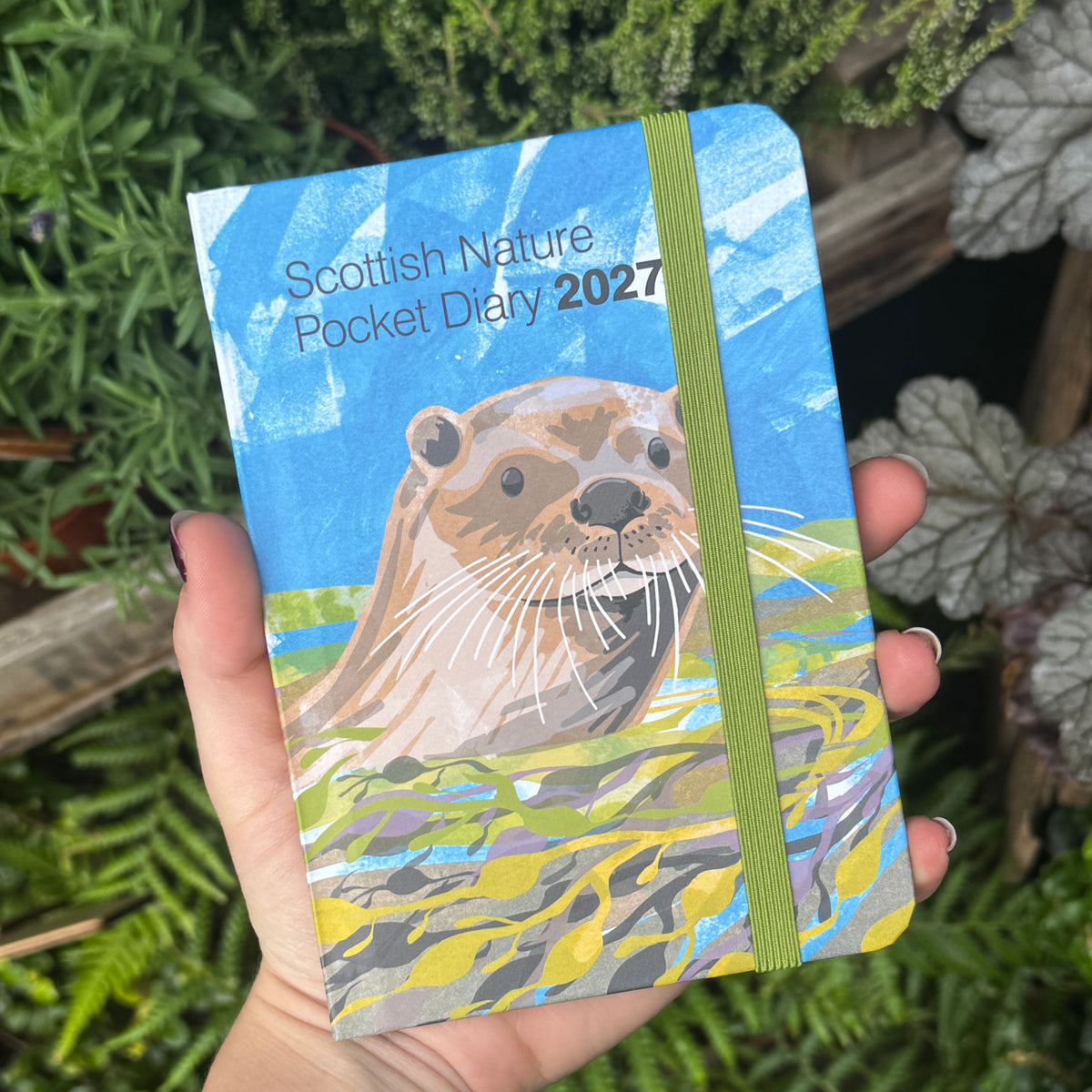 A hand holds a hardcover pocket diary titled 'Scottish Nature Pocket Diary 2027' featuring a painted otter illustration on the cover, with a green elastic closure band, against a background of green foliage.