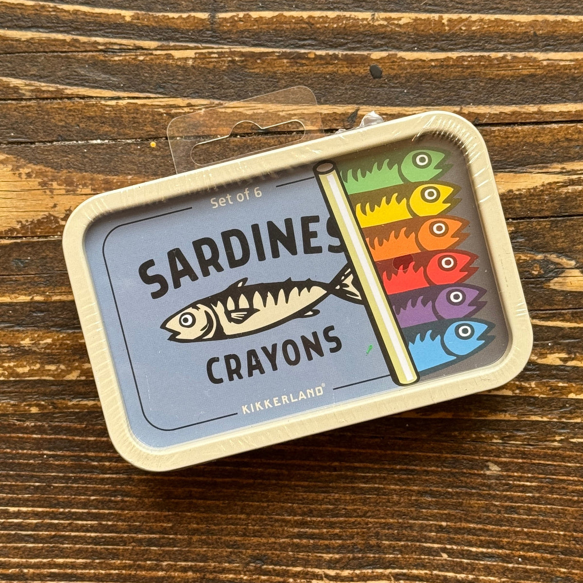 A Kikkerland Sardines Crayons set of 6 crayons in a tin package, placed on a rustic wooden surface. The packaging features a light blue background with a fish illustration and a vertical row of six colorful crayon tips.