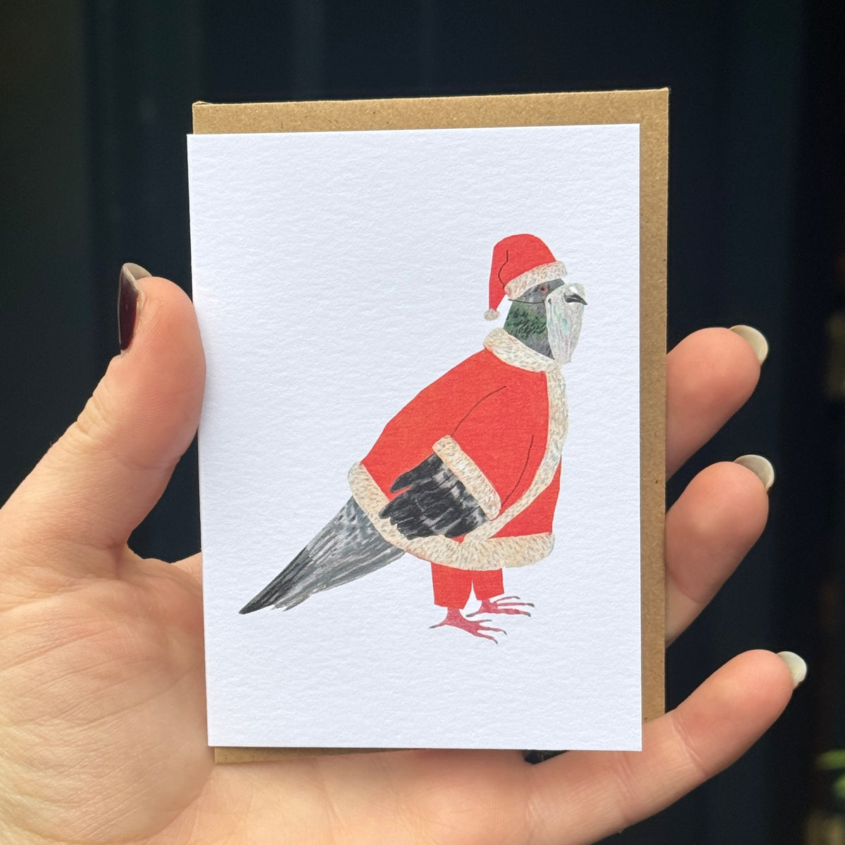 A hand holds a square greeting card featuring a painted bird dressed in a Santa hat and coat, with a brown envelope behind it. The background is a dark, softly blurred outdoor setting with yellow-green leaves on the left.